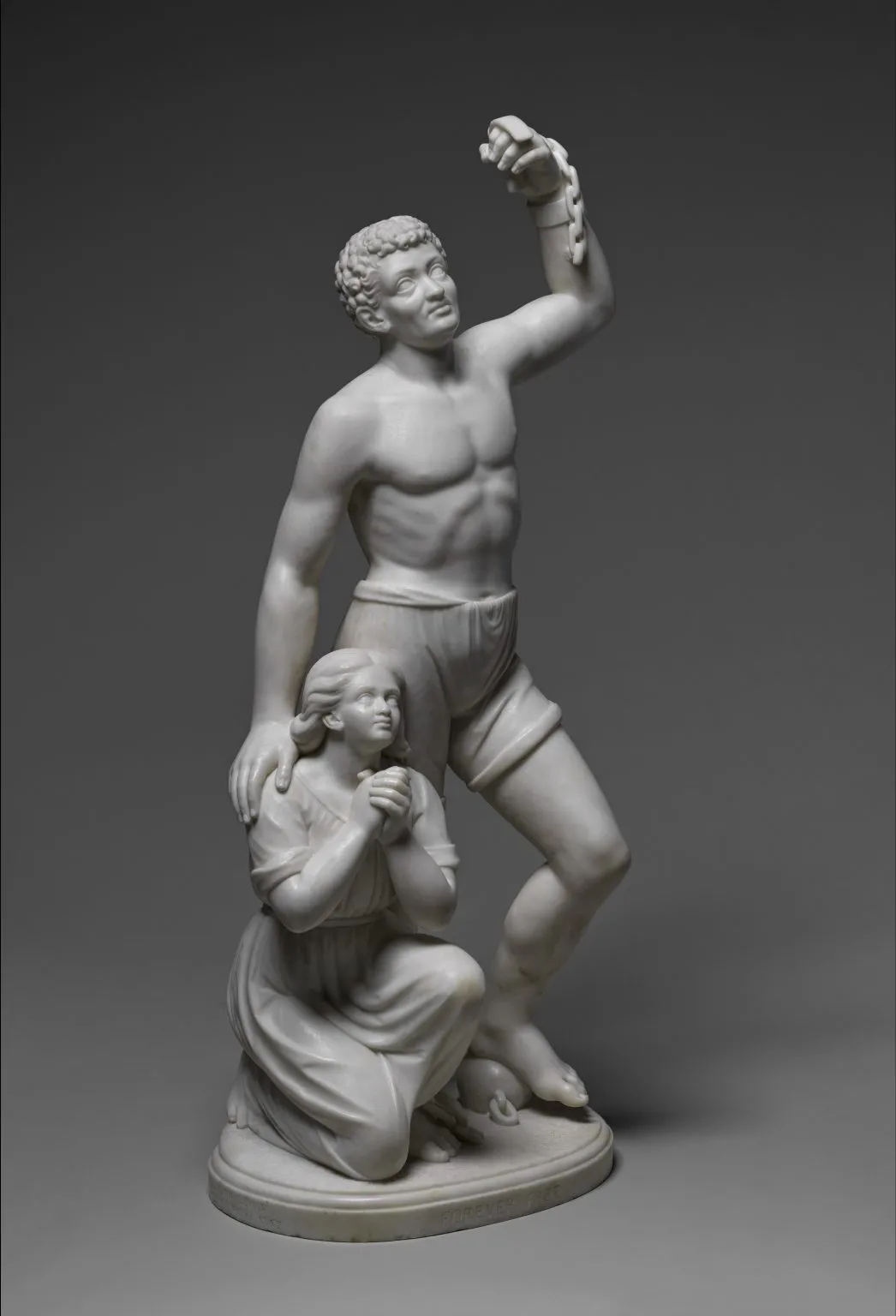 A photo of a white marble sculpture of two figures: a muscular young Black man standing with one arm raised and broken chains hanging from his wrist, and a kneeling young Black woman clasping her hands in prayer at his side, symbolizing emancipation.