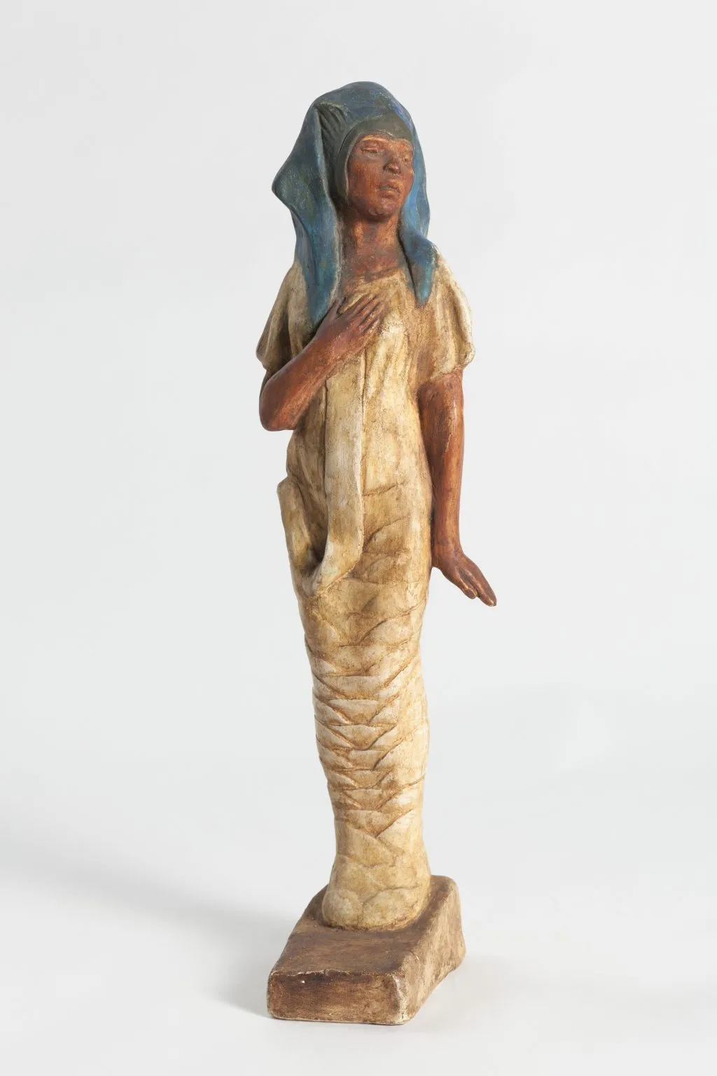 A photo of a small painted sculpture of a young Black woman with a serene expression, wearing a blue Egyptian pharoah-like head covering and a simple cream-colored dress, her right hand resting over her chest as her lower body tapers into a wrapped, column-like form.