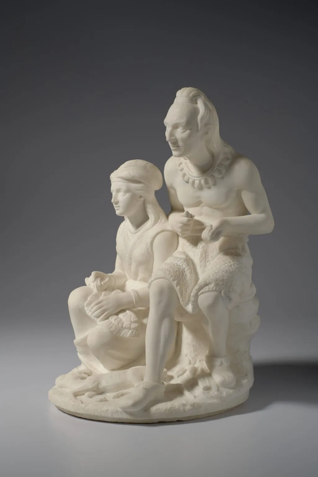 A photo of a white marble sculpture depicting two Native American figures: a middle-aged man with long hair and a necklace seated beside a young girl with braided hair, both in traditional clothing, shown in a calm, intimate pose.